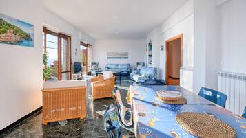 Ein geräumiges Wohnzimmer mit einem großen Esstisch, Korbmöbeln und Meerblick durch große Fenster.