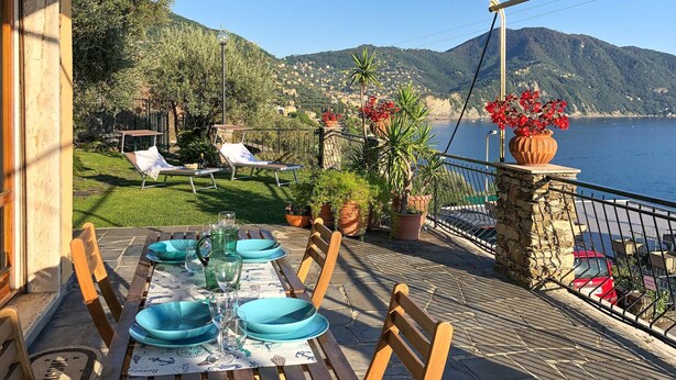 A terrace with a table set for dining, overlooking a coastal area with mountains in the background.