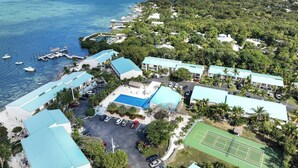An aerial view of a resort with multiple buildings, a swimming pool, tennis court, and boats docked near the water.