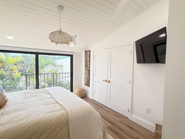 A bedroom with a bed, a ceiling fan, a TV mounted on the wall, and a sliding glass door leading to a balcony with plants.