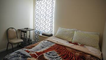 A bedroom with a bed, a chair, a small table, and a window with a patterned curtain.