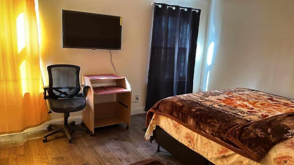 A bedroom with a bed, a desk with a chair, a TV mounted on the wall, and a window with curtains.
