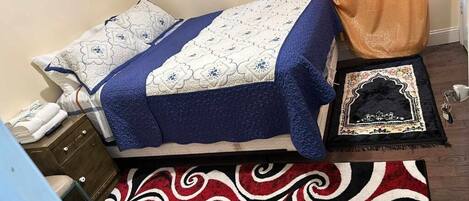 A bed with a blue and white quilt, a patterned pillow, a blue blanket, a decorative rug, and a small black and white rug with Arabic script.