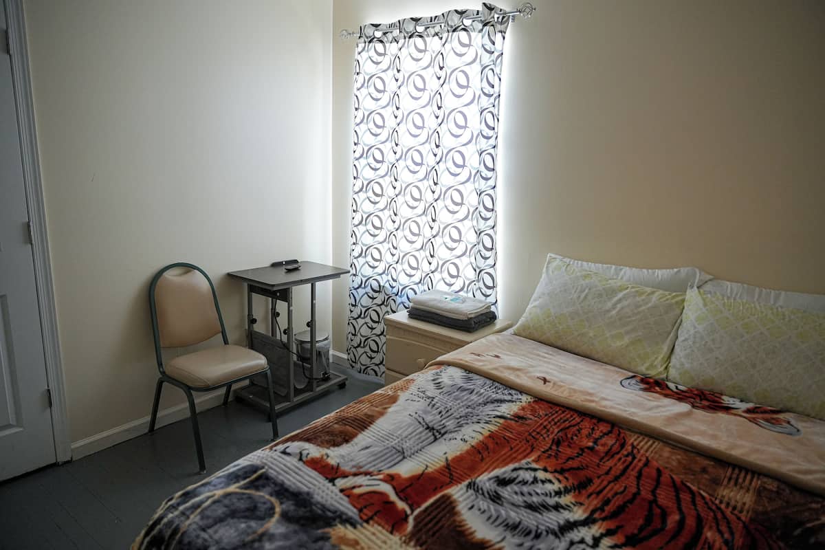 Ein Schlafzimmer mit einem Bett, einem Sessel, einem kleinen Tisch und einem Fenster mit gemusterten Vorhängen.
