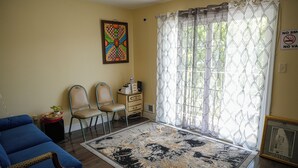 Ein Wohnzimmer mit einem blauen Sofa, zwei Sesseln, einem kleinen Tisch mit einer Pflanze, einem Teppich und einem Fenster mit Vorhängen.