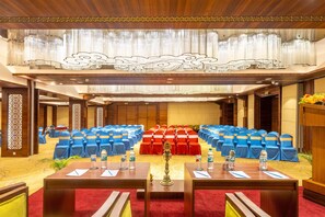 Un salón de conferencias con filas de sillas azules y rojas, una mesa central con botellas de agua y un diseño decorativo en el techo.