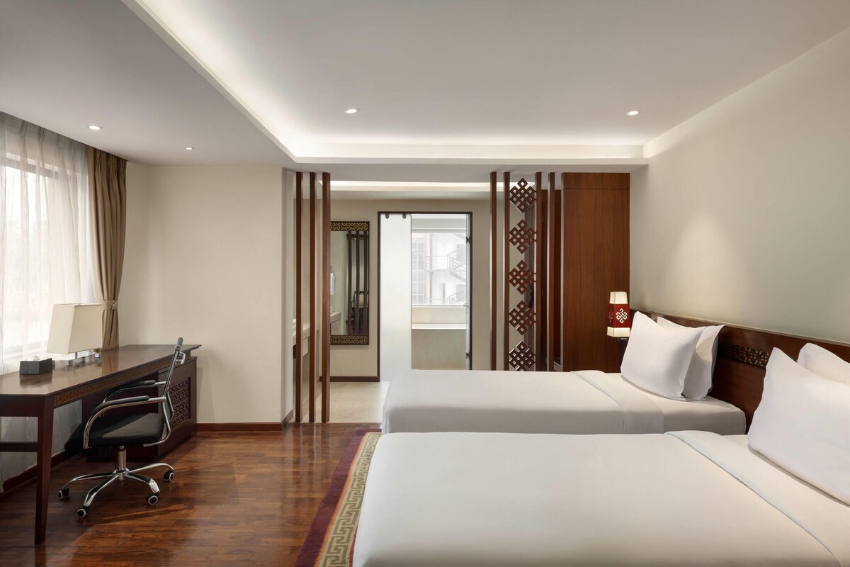 Habitació d'hotel amb dos llits, una taula de treball, una cadira, una làmpada i un panell decoratiu.