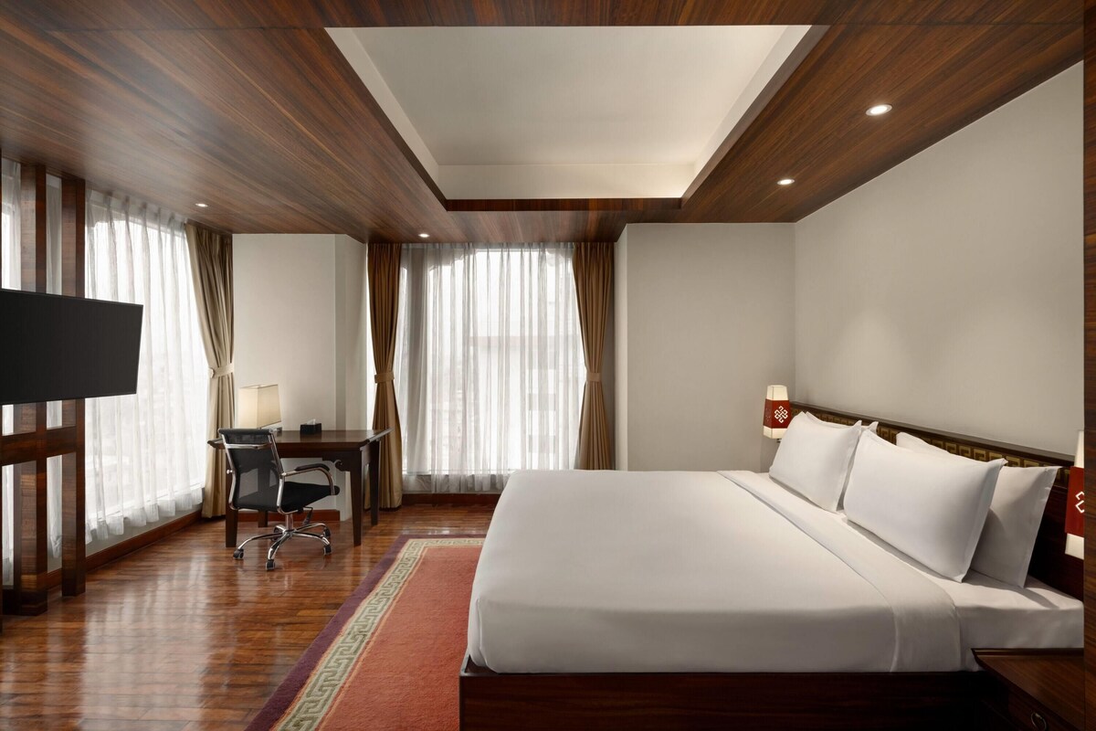 A hotel room with a wooden ceiling, a large bed, a desk with a chair, a TV, and a window with curtains.