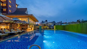 A swimming pool with lounge chairs and umbrellas, surrounded by a building with a tiled roof and a view of the city skyline. - Ramada by Wyndham Kathmandu Dhumbarahi (Kathmandu)