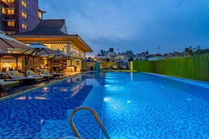 Una piscina con sillas de descanso y sombrillas, rodeada por un edificio con techo de tejas y vistas al horizonte de la ciudad.