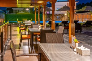 Un área de comedor al aire libre con mobiliario de mimbre, sombrillas y una piscina al fondo.