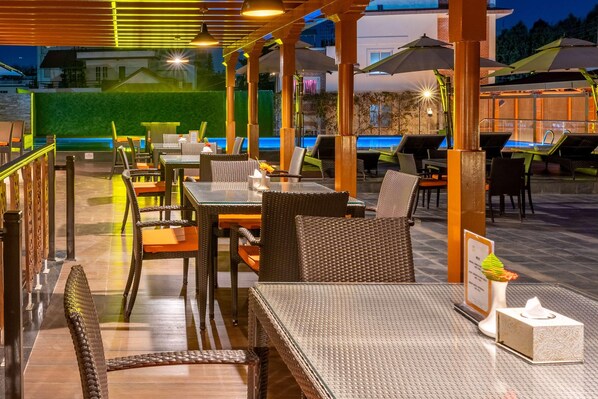 An outdoor dining area with wicker furniture, umbrellas, and a pool in the background. - Ramada by Wyndham Kathmandu Dhumbarahi (Kathmandu)