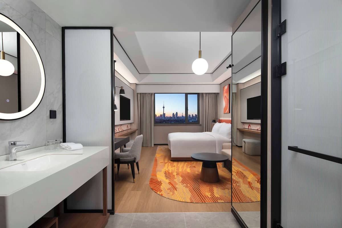 A modern hotel room with a large bed, a sitting area with a sofa and armchairs, a round coffee table, a large mirror, and a view of a cityscape through the window.