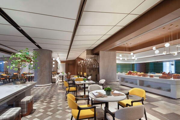Un comedor moderno con mesas redondas y sillas amarillas, una barra central con una larga barra, y una planta decorativa en el centro.