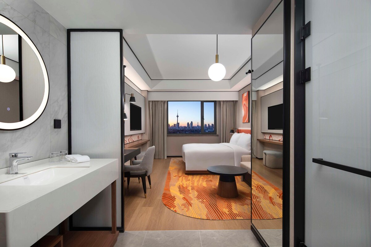 Habitación de hotel moderna con una cama grande, una zona de estar con una silla, una mesa de centro redonda, un espejo grande y vistas de un paisaje urbano a través de la ventana.
