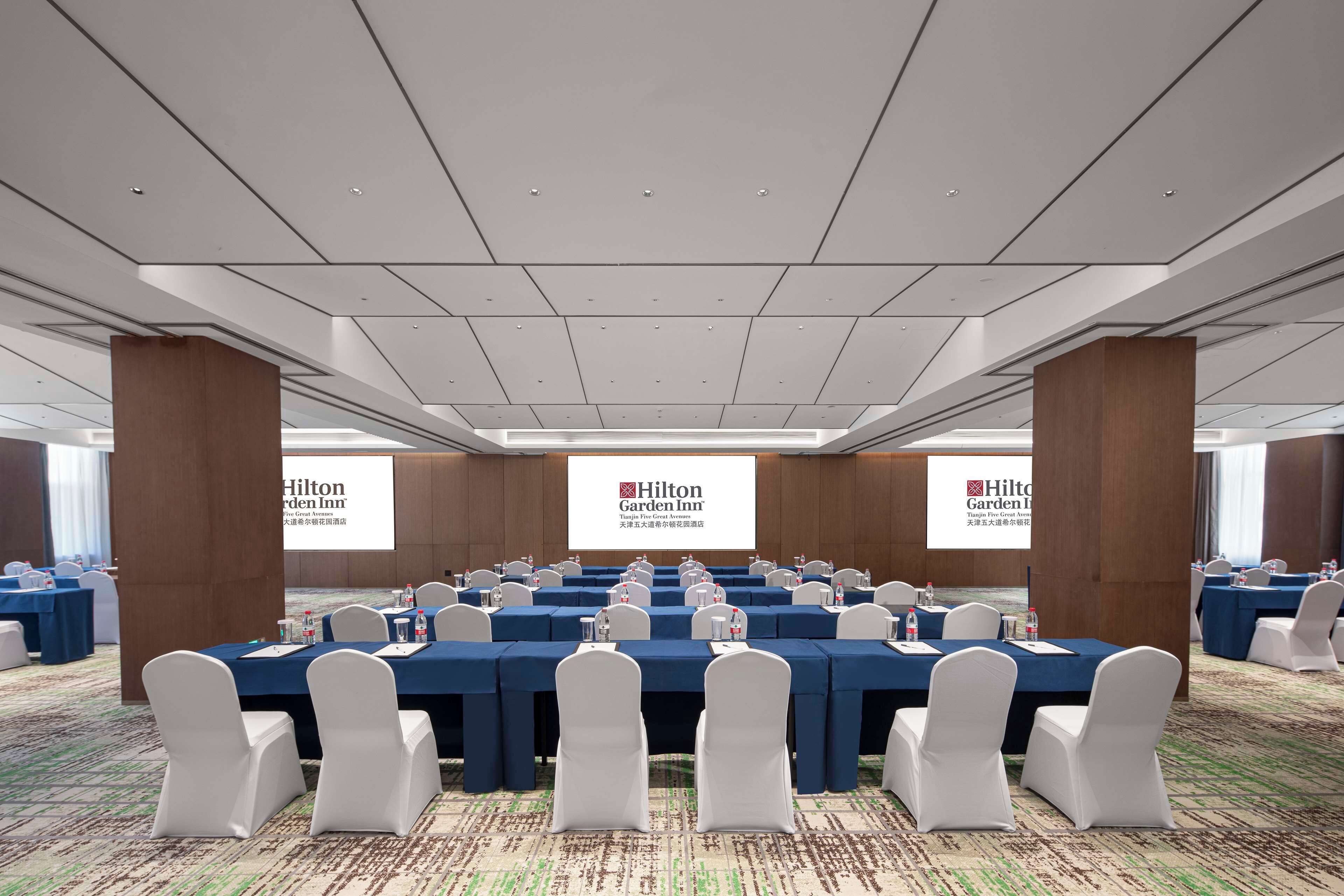 A conference room with multiple tables, white chairs, and a large screen displaying the Hilton Garden Inn logo.