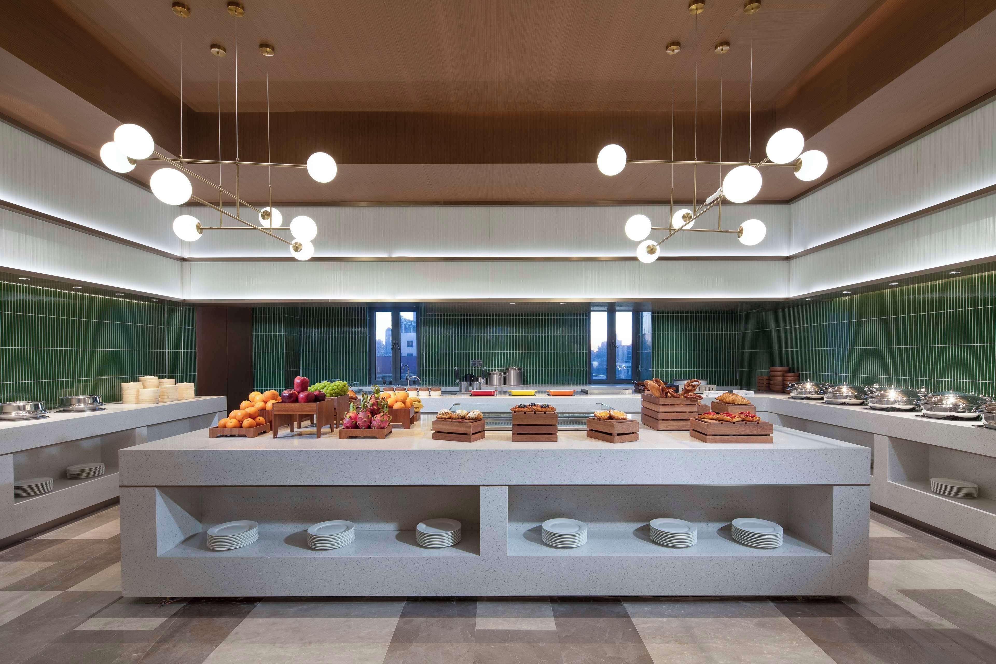 A modern breakfast bar with a variety of fruits and pastries, a clean and contemporary design with a green-tiled backsplash, and a ceiling with a unique lighting fixture.