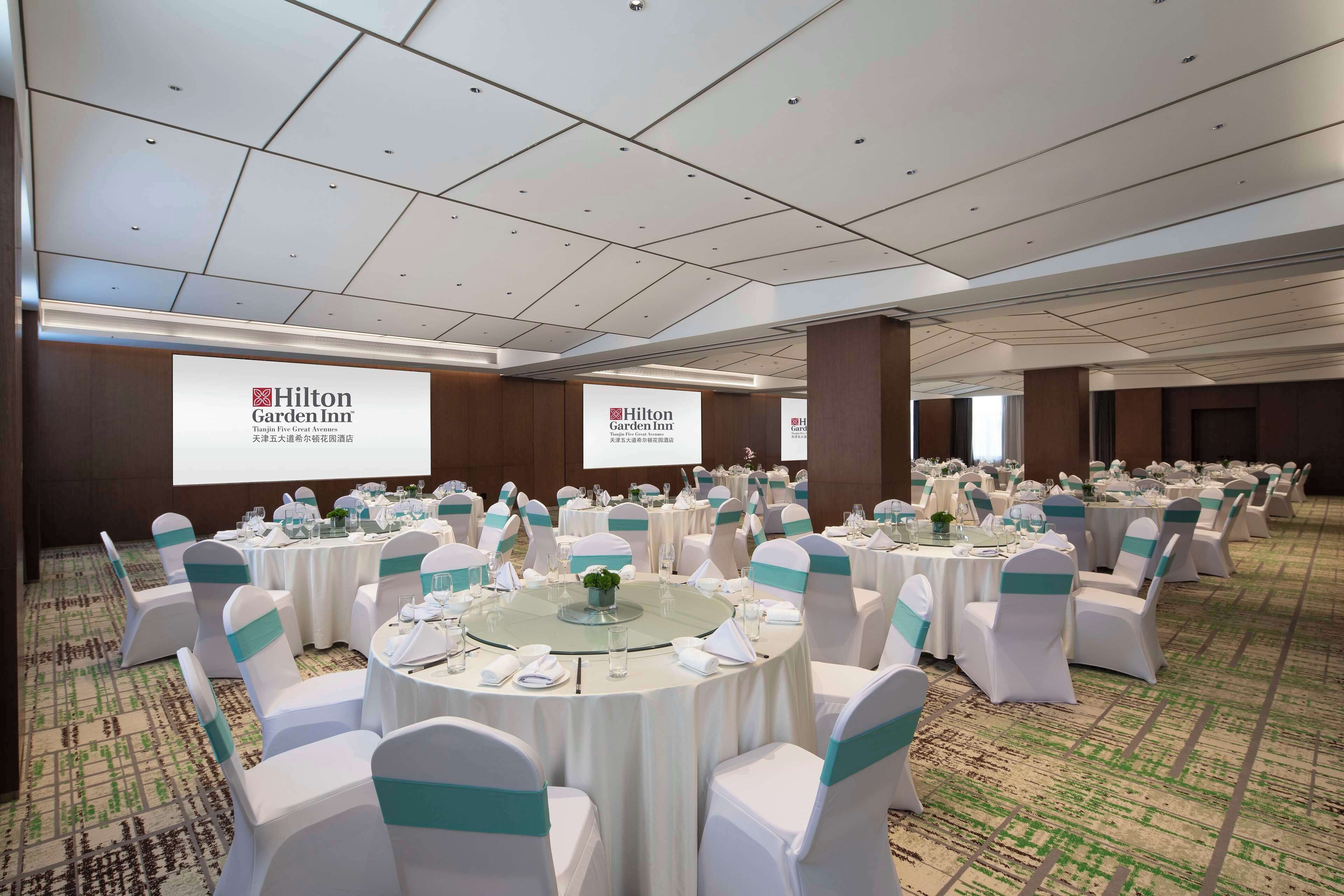 A large, well-lit conference room with round tables and white chairs, each with a teal sash. The room has a carpeted floor and a high ceiling with recessed lighting.