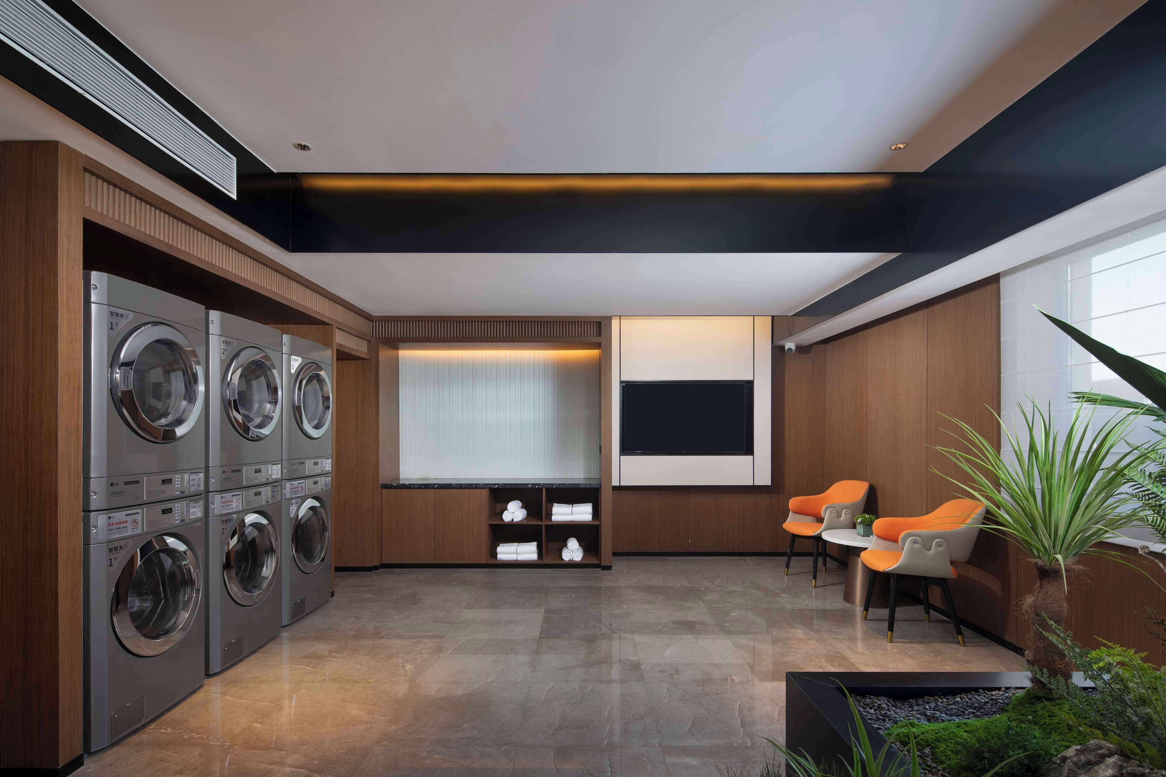 A laundry room with multiple washing machines, a seating area with chairs, and a large window with blinds.