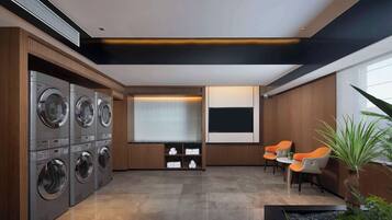A laundry room with multiple washing machines, a seating area with chairs, and a large window with blinds.