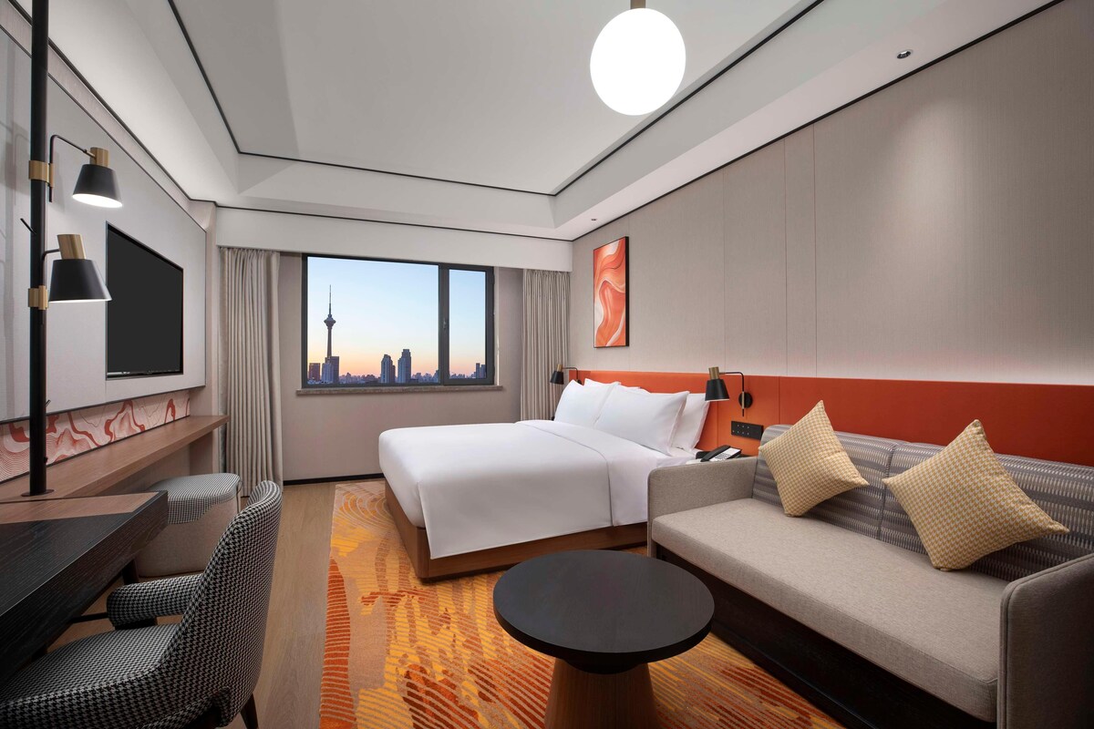 A hotel room with a bed, sofa, dining area, and a large window with a cityscape view.