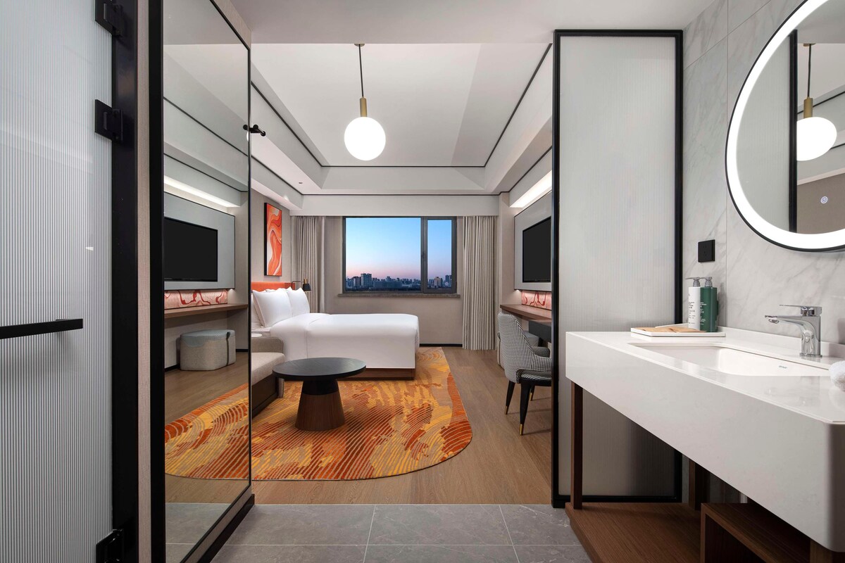 A modern hotel room with a large bed, a desk, a round mirror, and a view of the city through a large window.