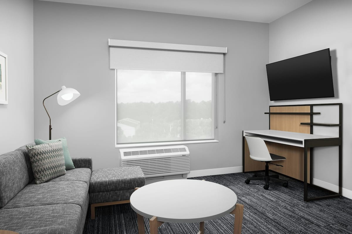 A modern hotel room with a gray sofa, a round white table, a desk with a chair, a mounted TV, and a large window with blinds.