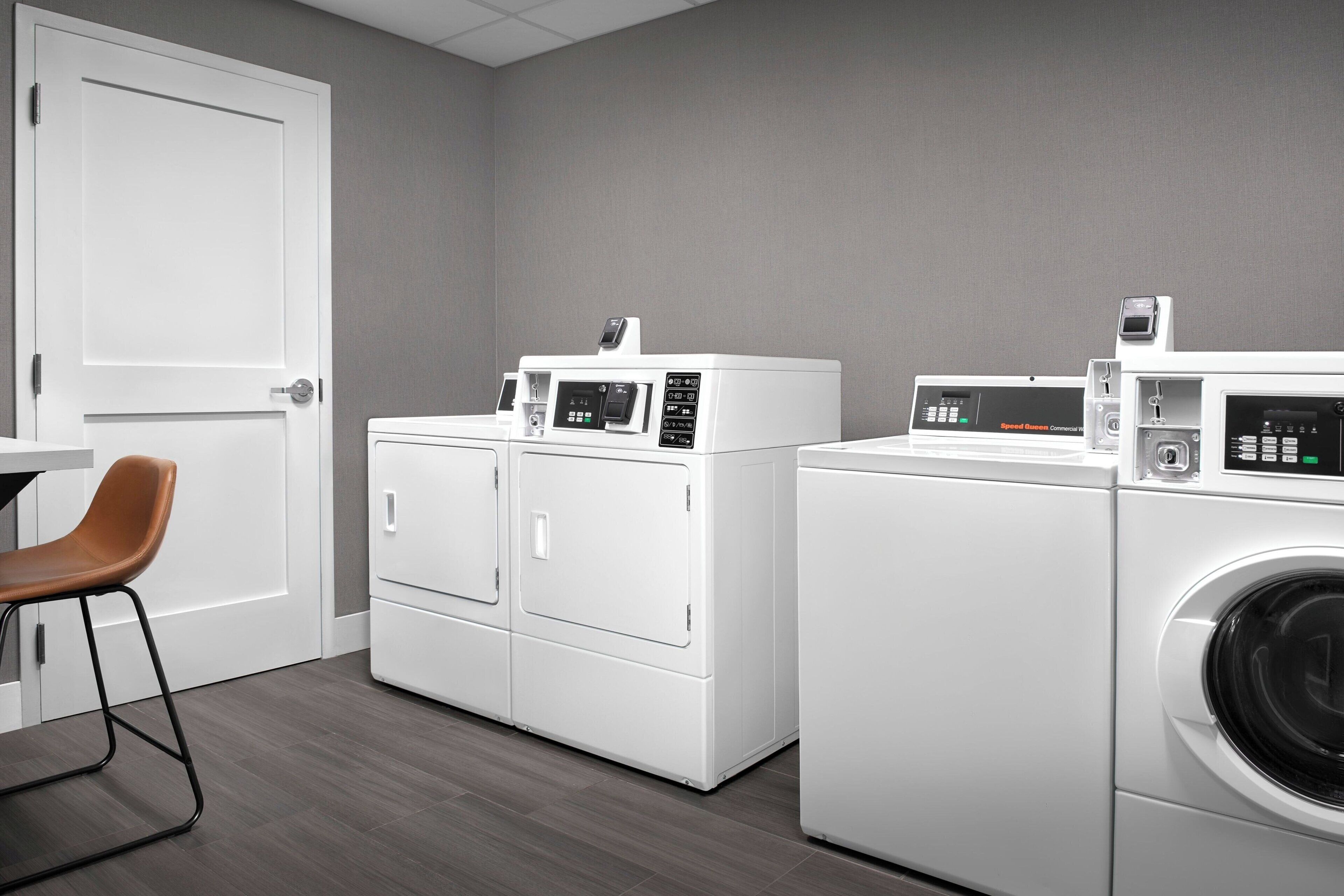 A laundry room with two front-loading washing machines and a dryer, a white door, and a brown chair.
