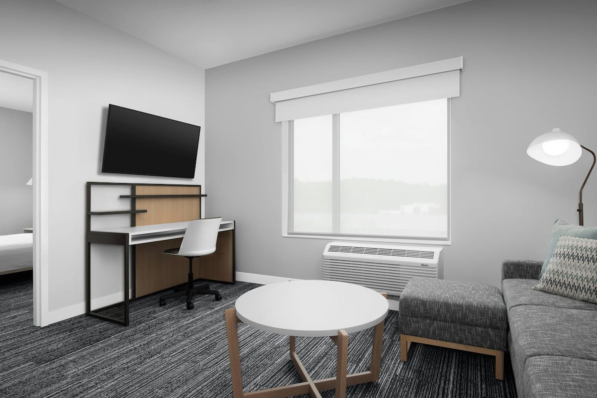 A modern hotel room with a flat-screen TV, a desk with a chair, a round table, a sofa, and a large window with a view.
