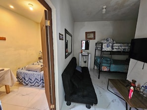 A compact room with a bunk bed, a small desk, a chair, a bedside table, a mirror, and a wall-mounted TV.