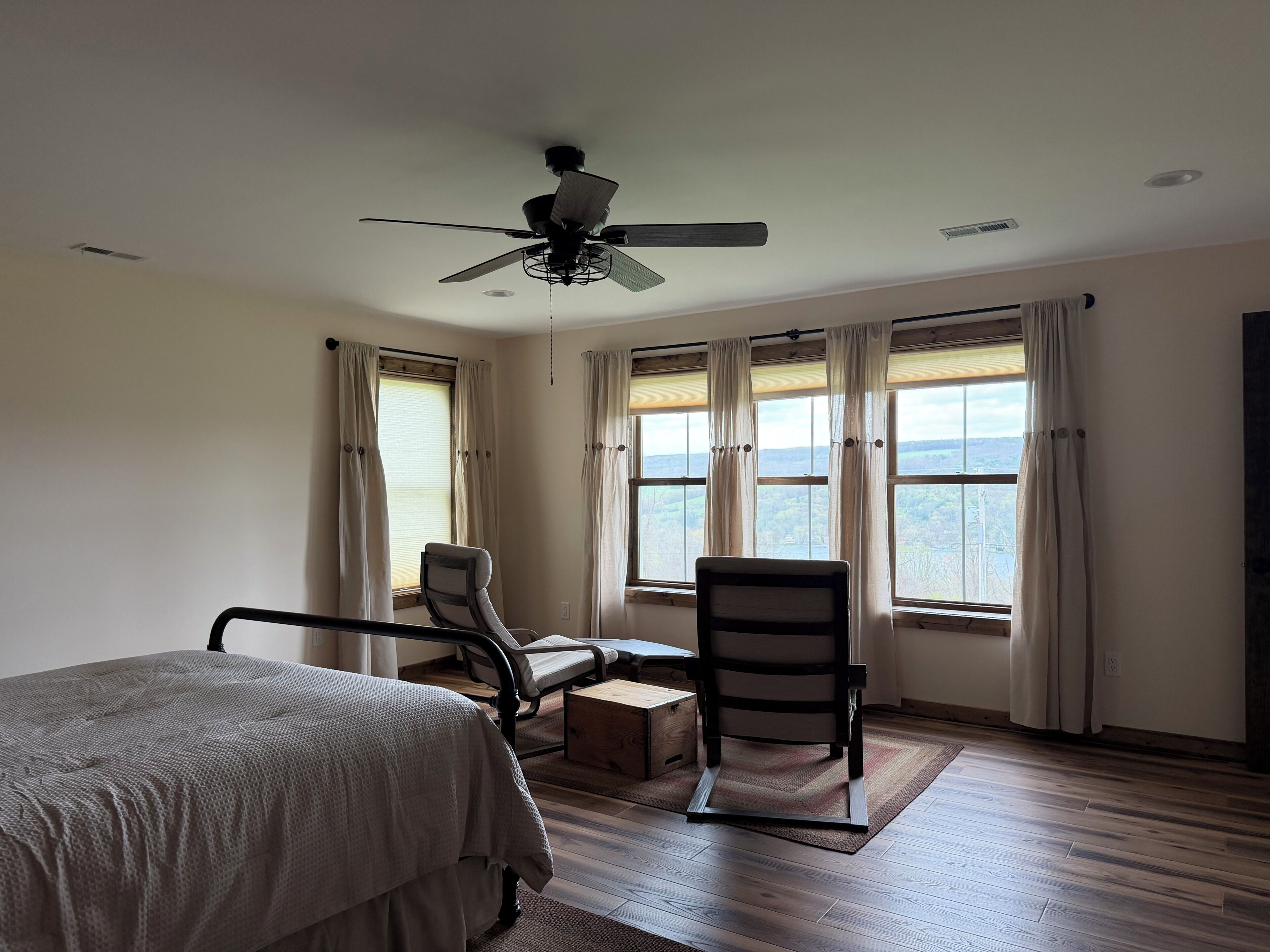 A bedroom with a bed, two chairs, a ceiling fan, and large windows with curtains.