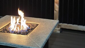 Ein Feuerkorb mit zwei Gläsern Rotwein auf einer Holzterrasse bei Nacht.