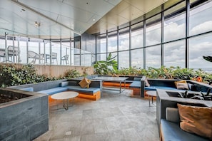 Un espace de détente extérieur moderne, doté de sièges, d’un foyer et de grandes fenêtres offrant une vue sur le ciel et les nuages.