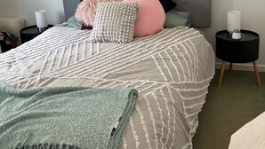 Een net opgemaakt bed met een grijze hoofdbord, meerdere kussens, een groene plaid en een nachtkastje met een lamp.