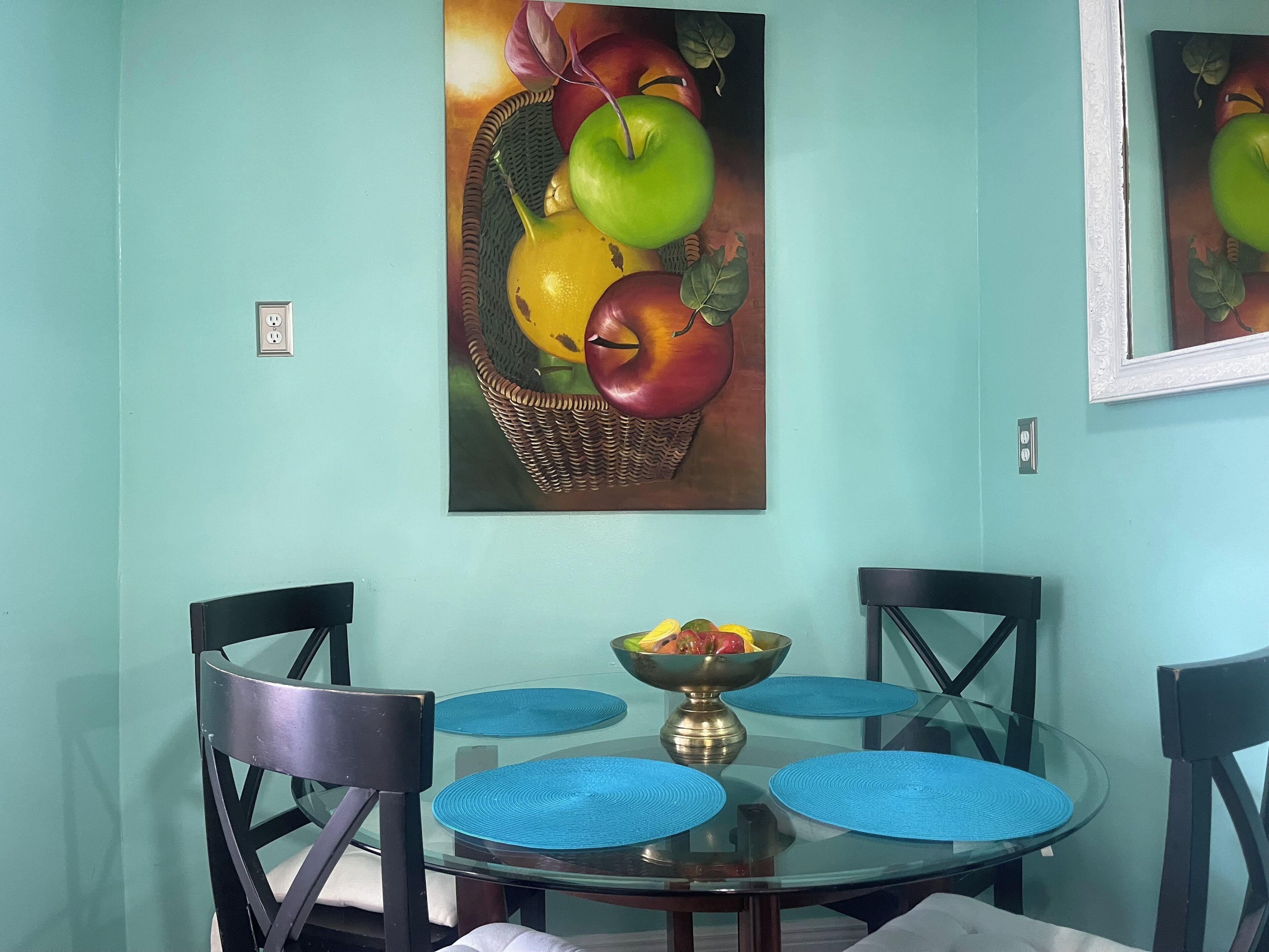 Uma área de refeições com mesa de vidro, jogos americanos azuis e cadeiras pretas. Há um quadro com uma cesta de frutas na parede.