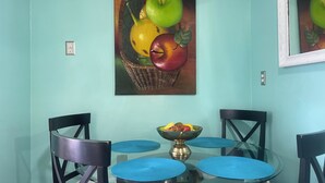 Un comedor con una mesa de vidrio, individuales azules y sillas negras. En la pared, un cuadro de una cesta de frutas.