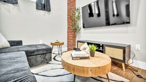 Una sala de estar moderna con un sofá gris, una mesa de centro redonda de madera, un televisor montado en la pared y un pilar de ladrillo.