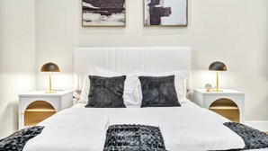 Una cama bien hecha con cabecera blanca, dos mesitas de noche con lámparas y dos cuadros abstractos en la pared.