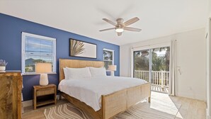 A bedroom with a bed, a ceiling fan, a window with blinds, a nightstand with a lamp, and a view of the outside balcony.