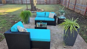 An outdoor patio area with black wicker furniture, including two sofas and a coffee table, all with teal cushions. There are two potted plants, one with long green leaves and another with a smaller, bushy plant. - The BlueStone-Stylish 3BR in Central Abilene (Abilene)