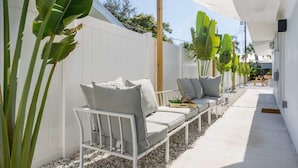 Eine Terrasse mit einer weißen Outdoor-Sofa, einem kleinen Tisch und einem Stuhl. Große, grüne Pflanzen und ein weißer Zaun begrenzen den Bereich.