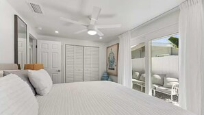 Ein Schlafzimmer mit einem großen Bett, einem Deckenventilator, weißen Wänden, einer Tür und Blick auf einen Balkon mit Sitzmöbeln.