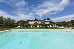 Una piscina con sillones de descanso y sombrillas, un edificio grande al fondo y un cielo despejado.