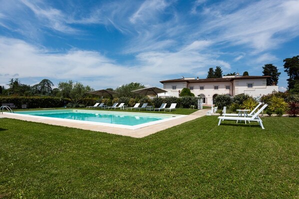 Una piscina con sillones de descanso, un césped cuidado y un edificio grande al fondo.