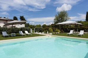 A swimming pool with lounge chairs and umbrellas, surrounded by greenery and a clear sky.