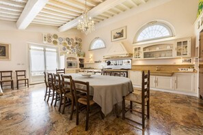 A spacious dining area with a large round table, wooden chairs, a chandelier, and a kitchen with white cabinets and a window.