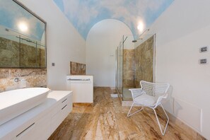 Un baño con un espejo grande, un lavamanos blanco, cabina de ducha de vidrio y una silla blanca.