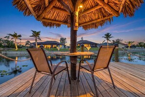A wooden deck with a thatched umbrella, two chairs, and a table with a glass. - Captivating Paradise, Pool Table, Tiki Hut, Kayak - Cape Sunset Villa - Roelens (Cape Coral)