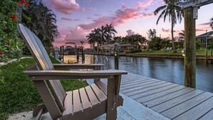 A wooden deck with a chair and a table, overlooking a calm water body with palm trees and a sunset sky. - Waterfront Living, Dock, Heated Pool & Spa - Gulf Gate Oasis - Roelens (Cape Coral)