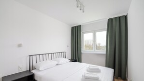 A neatly made bed with white linens and two pillows, a bedside table with a lamp, a window with green curtains, and a view of buildings outside. - Elbląska Modern Apartment (Warszawa)